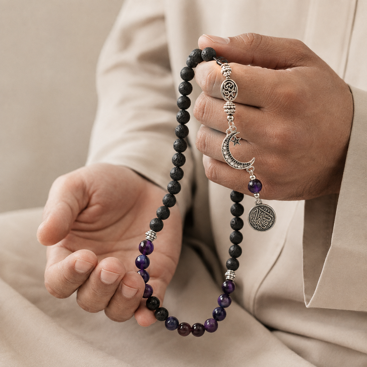 A close-up of a person holding a black and purple beaded prayer necklace with silver-toned crescent moon and circular charms, wearing a beige robe.