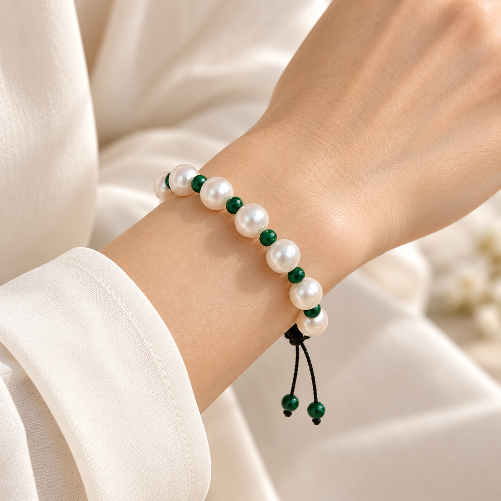 A close-up of a person's wrist wearing a beaded bracelet made of white pearls and dark green beads, with a black cord tassel, against a soft cream-colored fabric background.