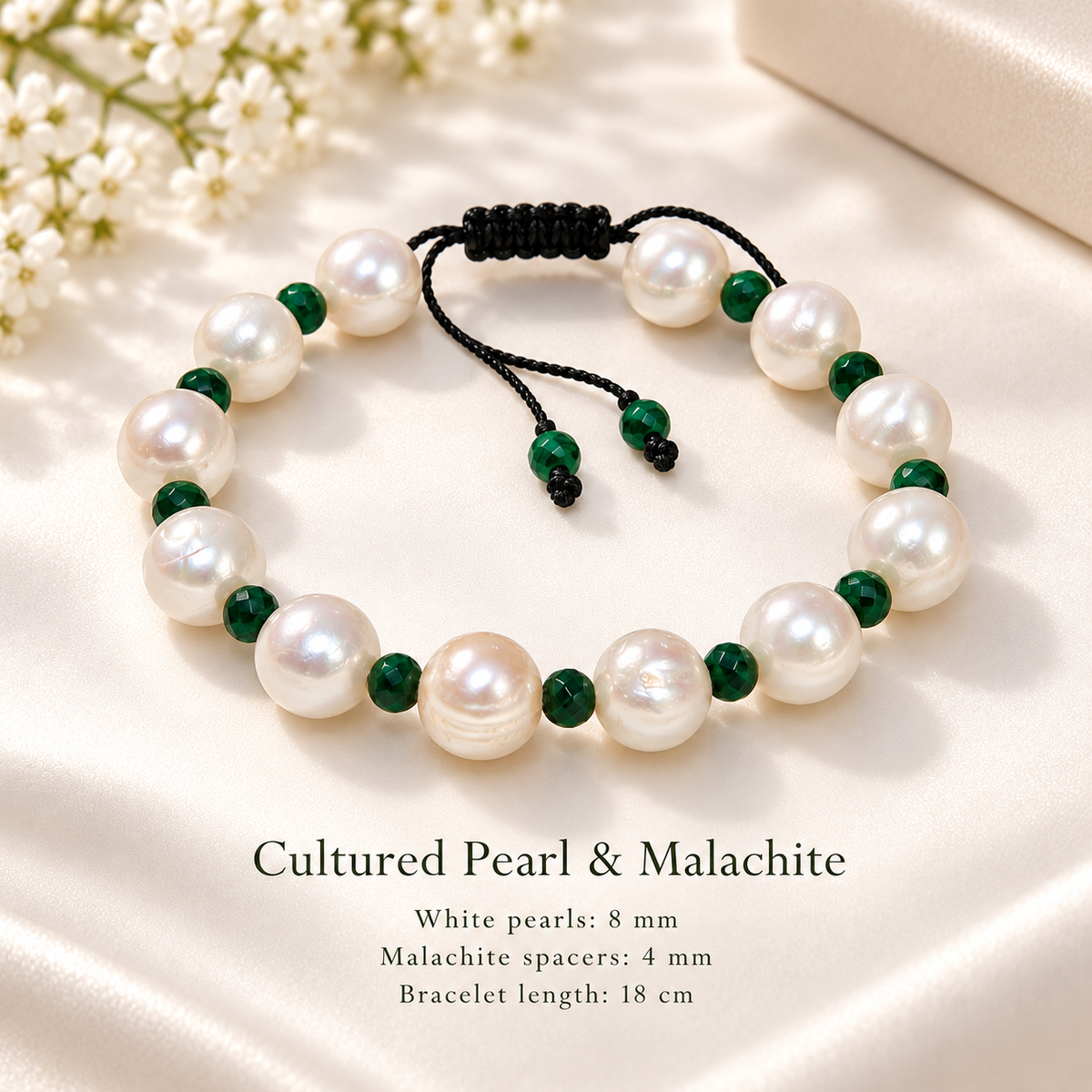 A pearl and malachite beaded bracelet displayed on a cream satin surface with small white flowers in the corner. The bracelet features large white pearls and dark green malachite beads separated by black cord.
