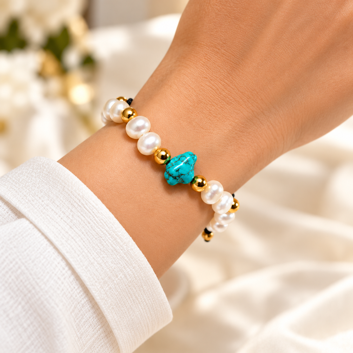 A close-up of a person's wrist wearing a beaded stretch bracelet with white faux pearls, small gold spacer beads, and a central turquoise-colored bead, set against a soft white fabric background.