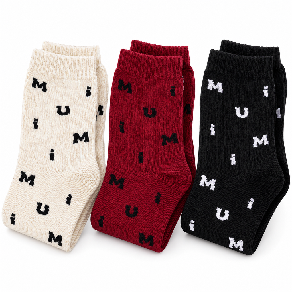 Three pairs of crew socks displayed side by side on a white background: one cream pair with black lettering, one dark red pair with black lettering, and one black pair with white lettering. The letters spell out 'MUM' repeatedly.