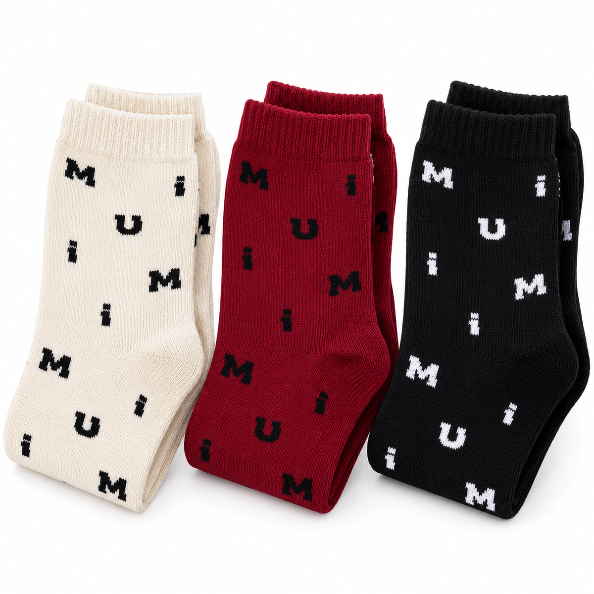 Three pairs of crew socks displayed side by side on a white background: one cream pair with black lettering, one dark red pair with black lettering, and one black pair with white lettering. The letters spell out 'MUM' repeatedly.