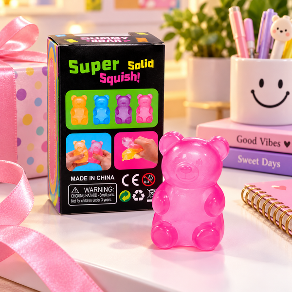 A pink translucent gummy bear toy sits in front of its black retail box on a tabletop. The box reads 'Super Solid Squish!' and shows colorful bear illustrations, with a pink ribbon, books, and a smiley-face pen cup in the softly blurred background.