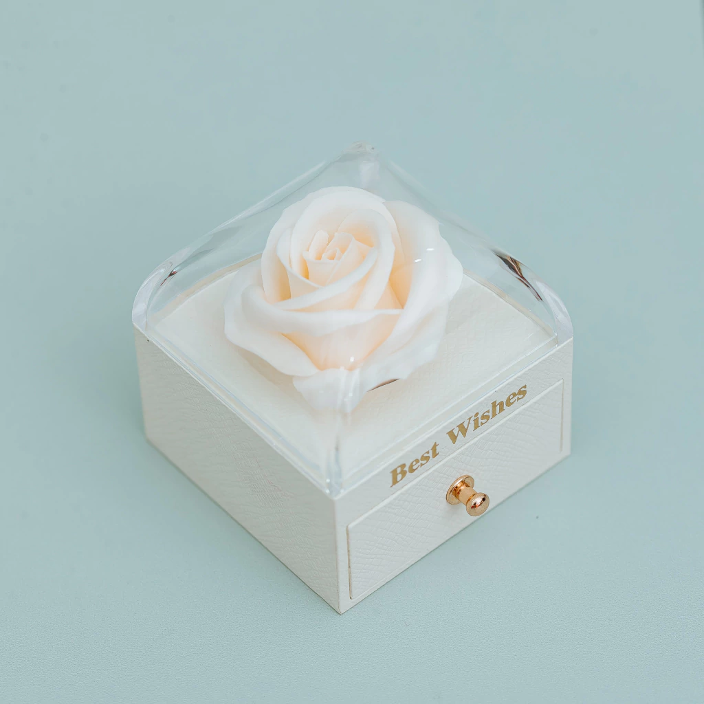 A small white gift box with a clear hinged lid, featuring a preserved white rose on a cream insert and gold 'Best Wishes' text on the front drawer. The box has a small gold pull and is photographed on a light blue background.