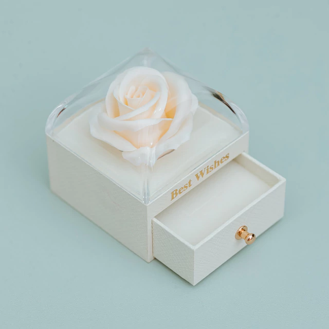 A cream-colored drawer box with a clear lid, featuring a preserved white rose on top and the words 'Best Wishes' printed on the side. The front drawer is pulled open, showing a small gold pull knob, set against a solid pale blue background.