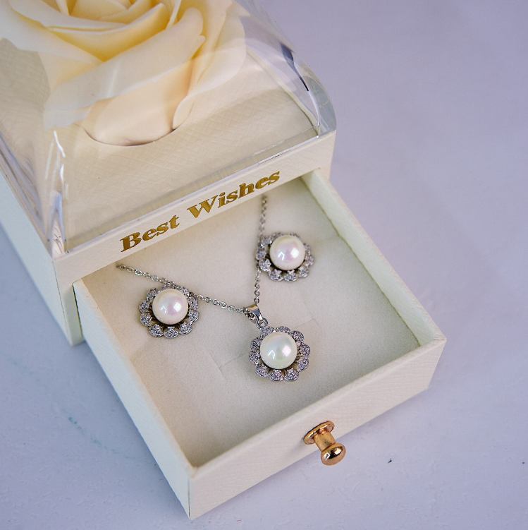 A pearl necklace and matching drop earrings displayed inside an open cream-colored jewelry gift box with a clear lid and a cream rose, on a light lavender background.
