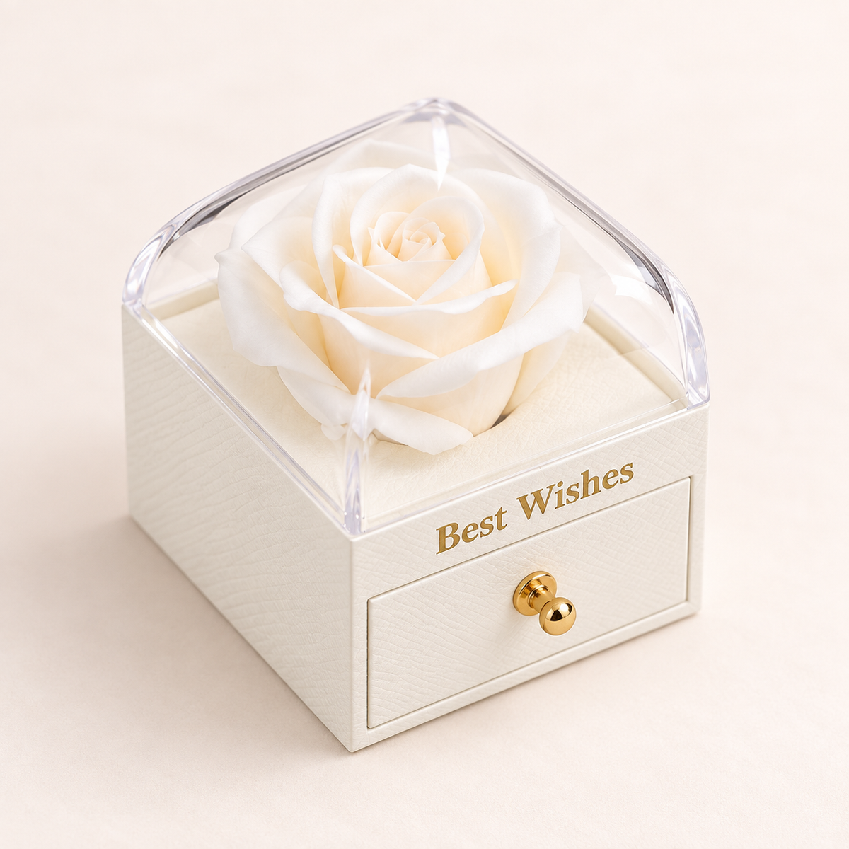 A preserved white rose displayed inside a small cream-colored square box with a clear lid and a single front drawer. The box is engraved with the words 'Best Wishes' in gold lettering and has a small gold knob.