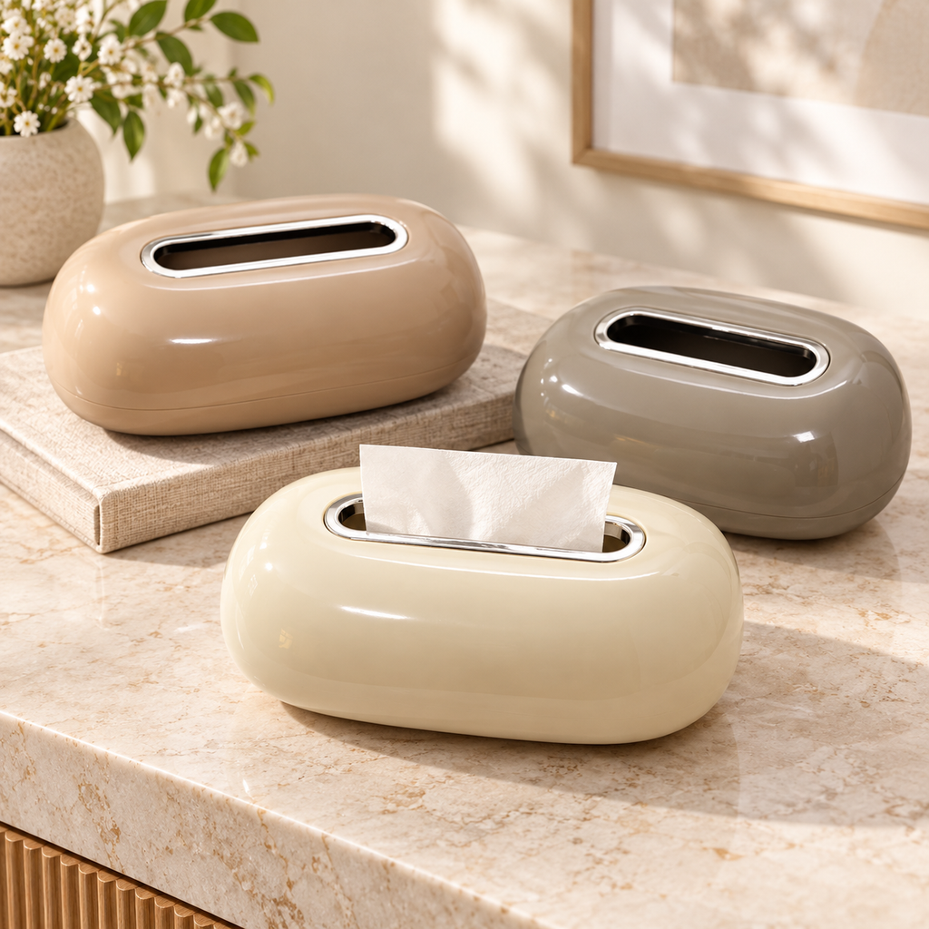 Three glossy oval tissue box dispensers in beige, taupe, and cream are arranged on a marble countertop with a vase of flowers and framed wall art in the background.