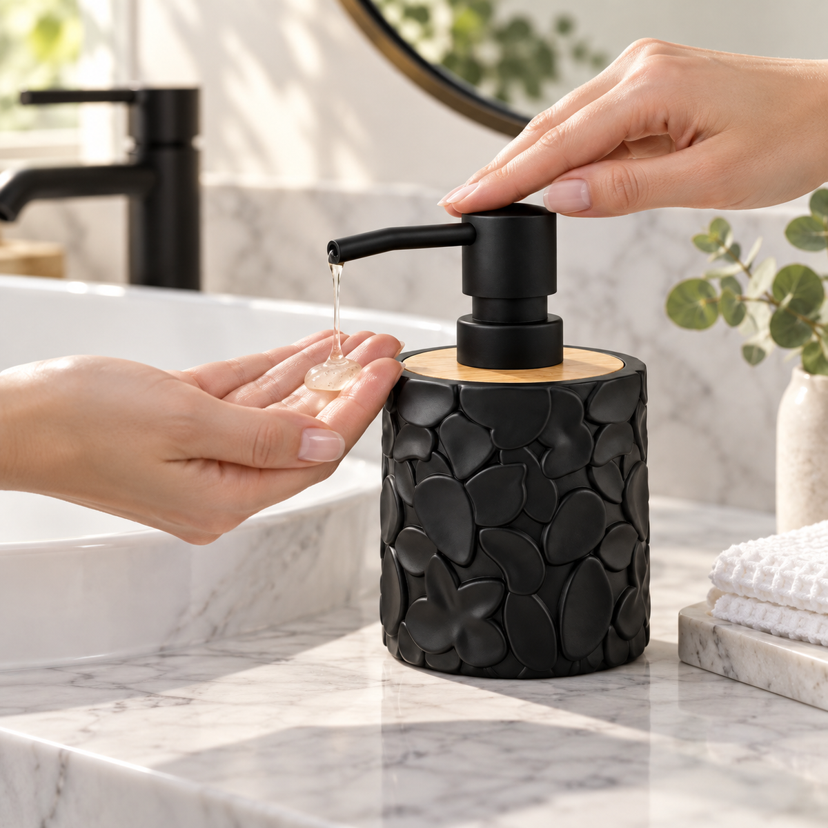 A black soap dispenser with a textured, raised petal-like pattern and a light wood top sits on a white marble countertop. A hand is shown pressing the black pump, with soap dripping into another hand.