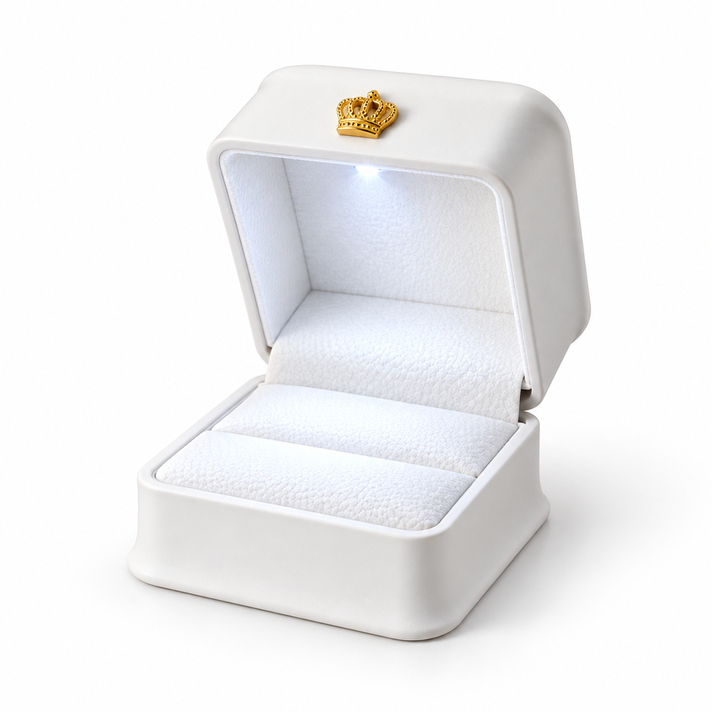 A white ring box with an open lid, featuring a small gold crown emblem on the inside of the lid. The box has a soft white textured interior and is shown on a clean white background.