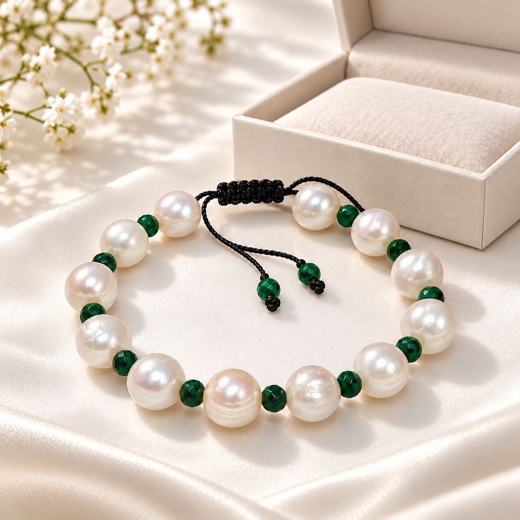 A beaded bracelet made of large white freshwater pearls and polished dark green beads, arranged on a cream satin surface with a cream jewelry box and small white flowers in the background.