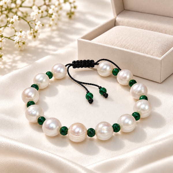 A beaded bracelet made of large white freshwater pearls and polished dark green beads, arranged on a cream satin surface with a cream jewelry box and small white flowers in the background.