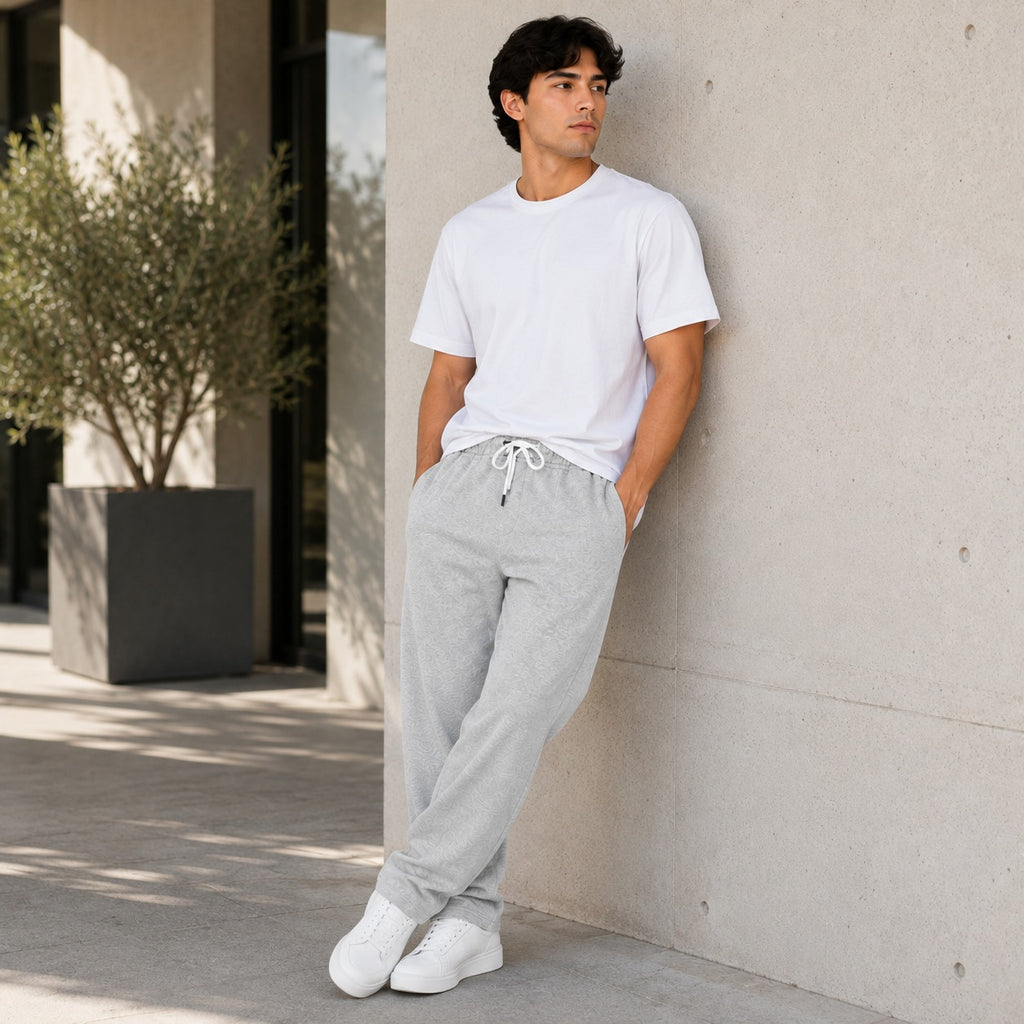 A man wearing a white crewneck t-shirt and light gray drawstring sweatpants leans against a concrete wall in an outdoor urban setting.